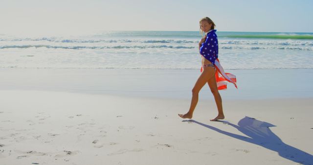 This image captures a woman walking on a sandy beach with an American flag draped over her shoulders. The ocean waves and bright sunlight create a serene and summery atmosphere. Ideal for use in marketing materials for travel and tourism, holiday celebrations, or patriotic-themed events. Also suitable for blog posts, social media, and advertisements focusing on freedom and American pride.