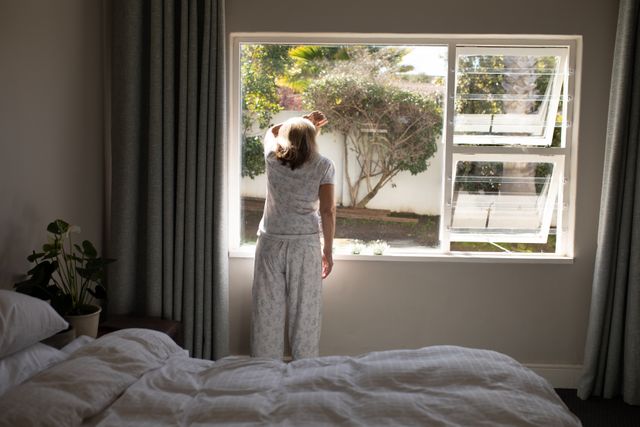 Senior woman wearing pajamas stands in her bedroom, looking out the window. The scene captures a peaceful morning moment at home, emphasizing themes of relaxation, solitude, and contemplation. Ideal for use in articles or advertisements related to elderly lifestyle, home living, quarantine during Covid-19, and mental well-being.