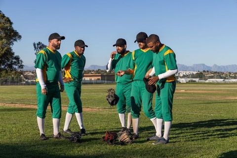 Baseball team in green uniforms huddling on sunny grass field with mitts and caps