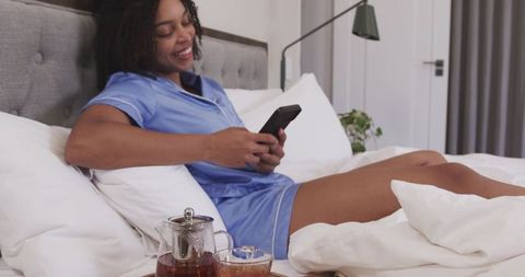 African american woman relaxing in blue satin pajamas on bed with tea and smartphone