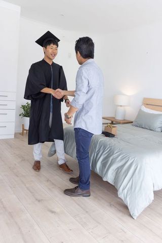 Young asian graduate celebrating shaking hands with friend in minimalist bedroom