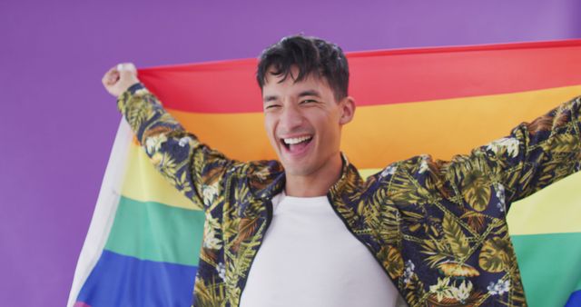Young person holding a vibrant rainbow flag and smiling against a purple background, symbolizing LGBTQ pride and the celebration of diversity and inclusion. Ideal for campaigns related to LGBTQ rights, pride month, community events, and social inclusion programs.