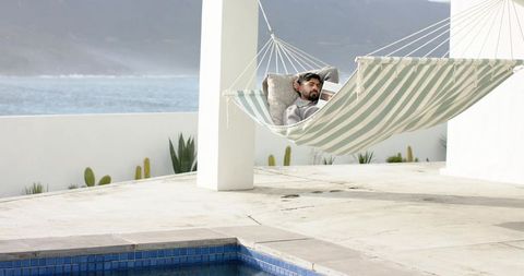Indian man reclining in striped hammock on coastal terrace with pool and sea view