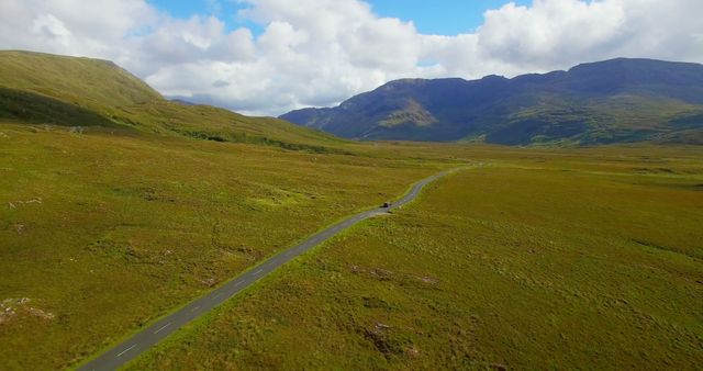 Long, winding road stretches through vast green Scottish Highlands, surrounded by lush hills and under partly cloudy sky. Use for promoting tourism, adventure travel, road trips, serene landscapes, nature exploration.