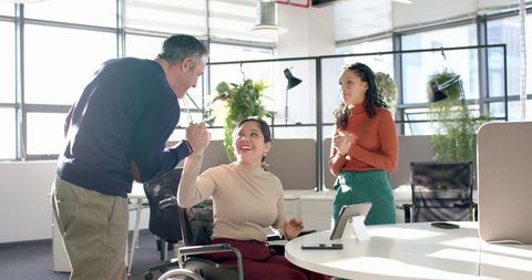 Colleagues fist-bumping with woman in wheelchair celebrating teamwork in open-plan office