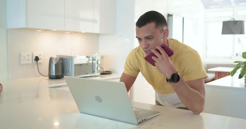 Young man leaning on marble island working on laptop and talking on smartphone in bright modern kitc