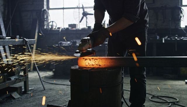 Grinding glowing steel billet with angle grinder in dark industrial workshop and sparks