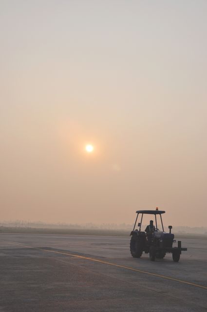 Tractor positioned on a foggy airport runway during sunrise creates a serene yet functional scene. Perfect for use in content about transportation, airport operations, early morning routines, and vehicles in airport settings.