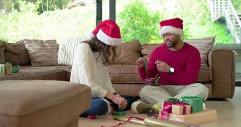 Couple wrapping christmas gifts on room floor wearing santa hats, cozy home holiday scene