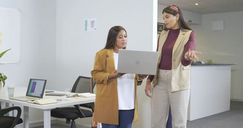 Diverse female coworkers collaborating over laptop in modern minimalist office workspace