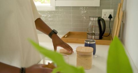 Man reaching to open wooden-lid jar on minimalist kitchen counter with plants and mug