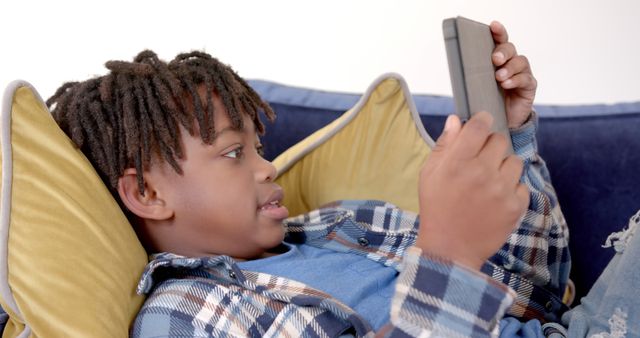 Young boy with dreadlocks is using a tablet while relaxing on a couch. The child appears immersed in the digital device, dressed in a blue shirt and plaid outerwear, exuding a sense of comfort. This image can be used for topics related to children's technology use, e-learning, digital entertainment, and family relaxation at home.