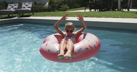 Young girl floating on pink donut pool float in sunny backyard wearing sunglasses