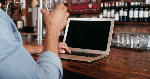 Person using laptop in bar while holding beverage, shows casual work environment and remote working lifestyle. Could be used to illustrate freelancing, modern workspace, technology in everyday life, or promoting work-life balance.
