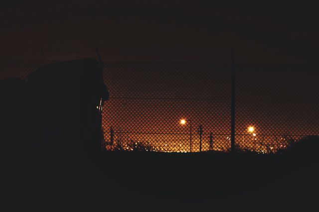 Cityscape scene captured at night with distant lights glowing behind chain link fence. Ideal for use in projects related to urban environments, night scenes, city safety, security themes, and contrasting light and dark visuals.