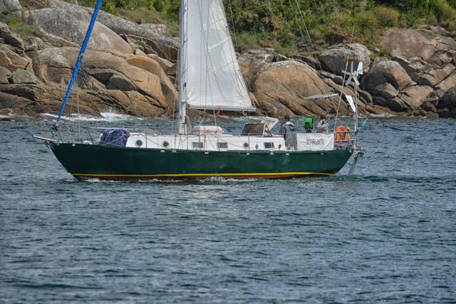 Green sailboat navigating nearby rocky coastal waters with calm blue ocean and scattered boulders. Ideal for illustrating concepts related to boating, maritime adventures, coastal travel, oceanic explorations, and marine transportation.