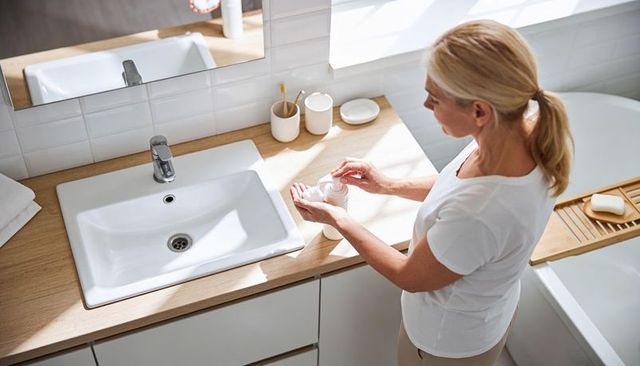 Mature woman dispensing face cream at sunlit wooden bathroom vanity with sink and bathtub