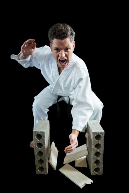 Karate practitioner in white uniform and black belt breaking wooden plank with hand against black background. Ideal for use in martial arts promotions, strength and determination concepts, sports training materials, and self-defense advertisements.