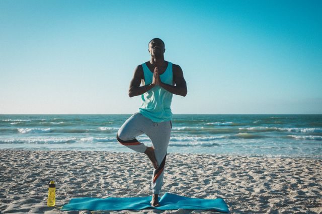 African American man practicing yoga on beach at sunset, standing on one leg in tree pose. Ideal for promoting fitness, healthy lifestyle, mindfulness, and outdoor activities. Perfect for wellness blogs, fitness websites, and meditation apps.