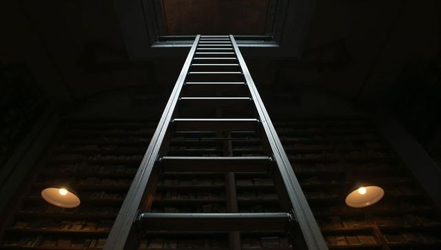Ascending metal ladder reaching ceiling hatch in dim atmospheric library between shelves