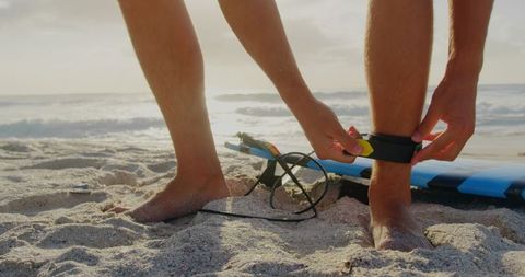 Person securing surfboard leash on sandy beach