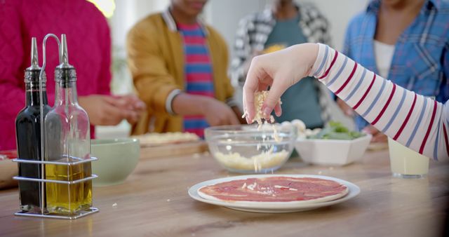This image shows a group of friends preparing a pizza together in a kitchen. One person is sprinkling cheese on a pizza base, while others are gathered around a bowl of ingredients. This can be used to depict themes of cooking, social gatherings, teamwork in the kitchen, homemade meals, or casual lifestyle. Great for advertising culinary classes, food blogs, or social events.
