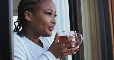 African american woman sipping tea on balcony in robe, looking out, morning calm