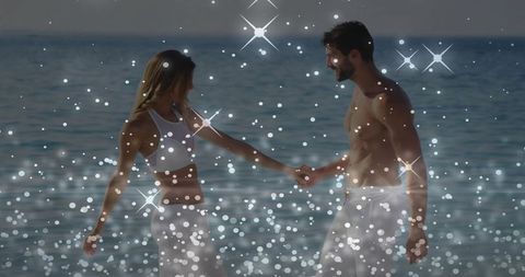Holding hands in sparkling seaside glow, couple walking waist-deep in ocean at sunset