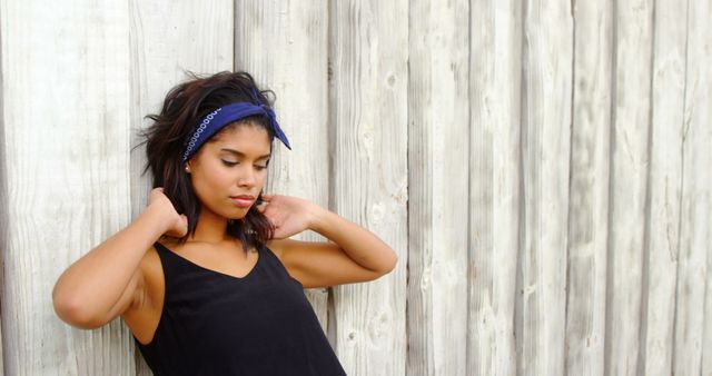Young woman wearing black top and blue bandana leaning against wooden wall outdoors. Perfect for lifestyle, fashion, casual wear, and relaxation themes. Great for blogs, social media posts, and advertising materials.