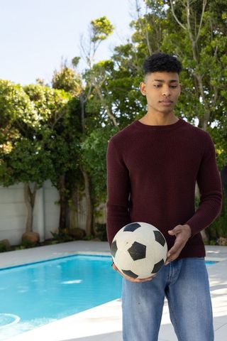African american teen standing poolside holding soccer ball in casual sweater and jeans