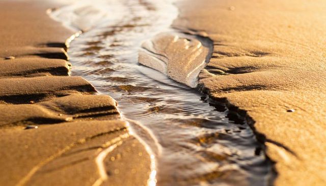 Flowing tidal rivulet carving golden sand with glinting ripples at golden hour closeup
