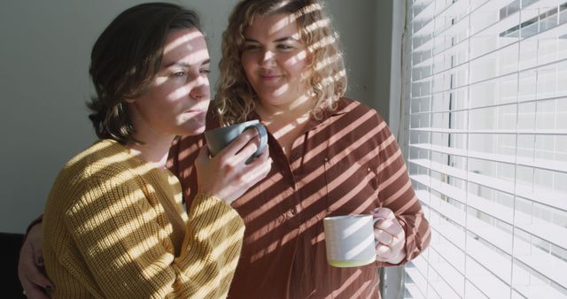 Same-sex couple enjoying morning coffee beside a sunlit window with blinds. Perfect for themes of love, togetherness, equality, home life, and casual morning routines. Ideal for LGBTQ representation in lifestyle imagery, domestic settings advertisements, and articles about relationships and morning rituals.