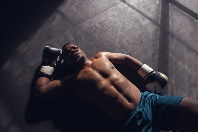 High angle view of tired shirtless african american young male boxer lying on floor in health club. Glove, exhausted, practicing, resting, unaltered, boxing, sport, training, strength and fitness.