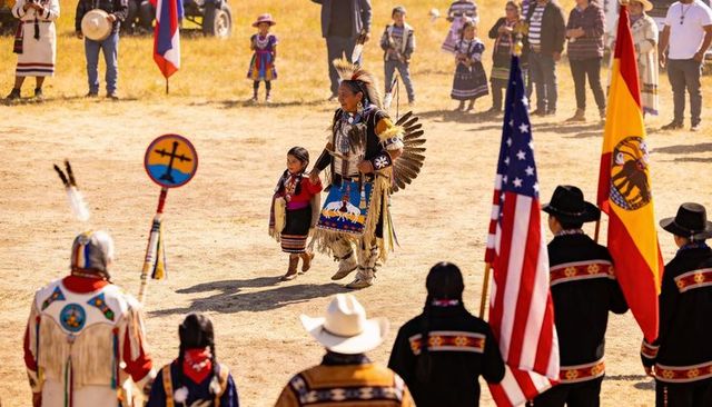 Walking dancer holding child's hand in powwow arena wearing feathered headdress and regalia