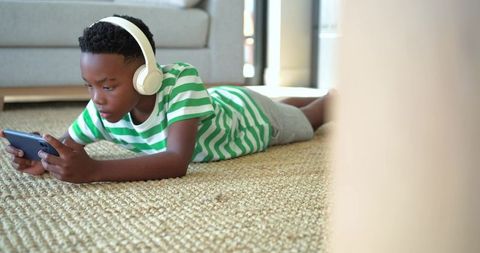 African american boy lying on rug using smartphone wearing headphones in cozy living room