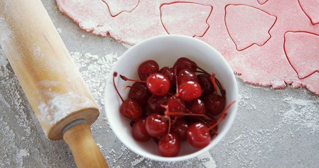 Perfect for illustrating recipes, baking guides, or culinary blogs, highlighting any dessert preparation process. This image showcases the vibrant red cherries in a bowl, a rolling pin dusted with flour, and heart-shaped dough cutouts. Ideal for enhancing content related to homemade baking, pie making, and dessert decoration.