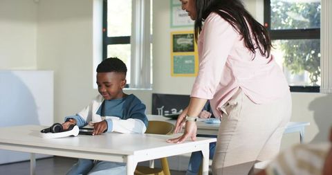 African American teacher guiding male student using tablet and headphones in classroom