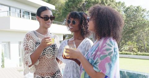 African american women enjoying refreshing drinks on modern pool deck by contemporary home