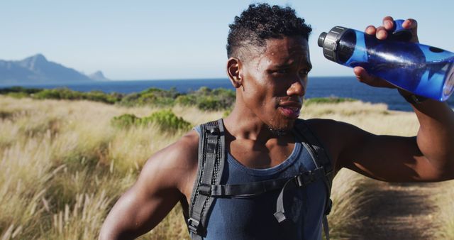 Young, fit man is taking a moment to hydrate with a sports bottle during an outdoor hike. He is surrounded by scenic views of mountains and the ocean. Perfect for materials related to fitness, active lifestyle, hydration, outdoor adventures, and summer activities.