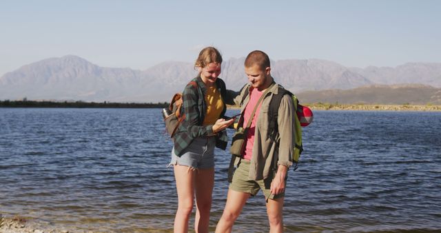 Young male and female hikers using smartphone beside a serene lake with mountains in the background. Ideal for articles promoting outdoor activities, technology's role in nature exploration, or showcasing healthy lifestyle and travel content.