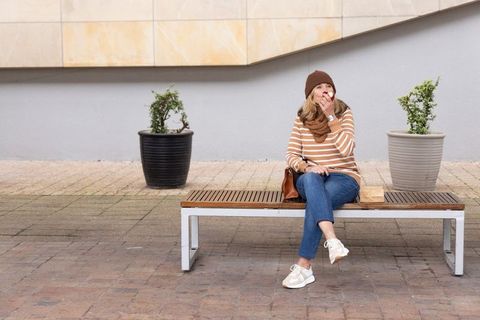 Woman enjoying apple while sitting on bench in urban plaza with planters and handbag