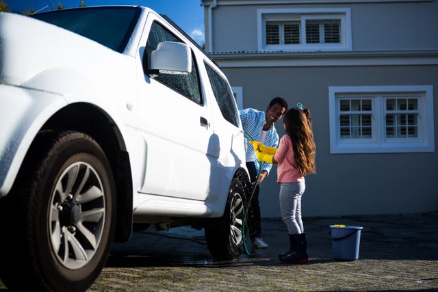 Father and daughter washing car together outside house. They are using a sponge and a bucket of soapy water. The scene depicts family bonding, teamwork, and outdoor fun on a sunny day. Ideal for use in advertisements, parenting blogs, family activity articles, and home improvement content.
