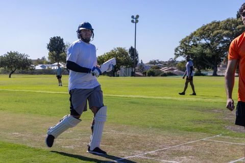 Batsman running along crease wearing helmet and pads on sunny park cricket pitch midplay