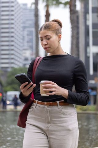 African american professional woman checking smartphone while holding coffee in downtown plaza