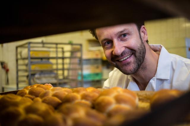 Baker smiling while removing freshly baked buns from oven. Ideal for content related to baking, culinary arts, food industry, and small business promotions. Perfect for illustrating the joy and skill involved in baking.
