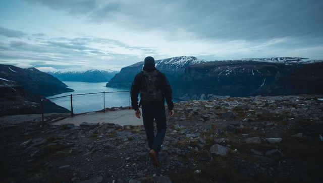 Solitary hiker walking toward fenced lookout over dramatic fjord and snowcapped mountains