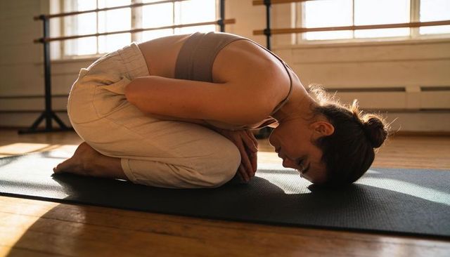 Restorative child's pose practicing on yoga mat in sunlit dance studio with barre