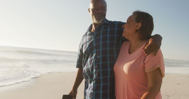 Elderly couple walking along sandy beach during daytime, smiling and looking at each other fondly. Ideal for campaigns promoting retirement, togetherness, lifestyle, or travel destinations.