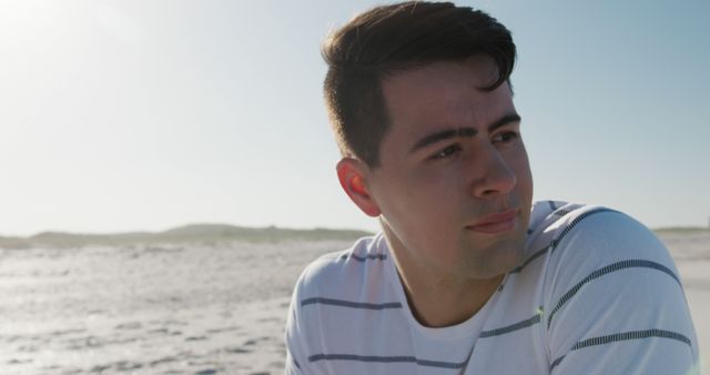 Young man wearing a striped shirt sitting on a sandy beach looking into the distance. Clear sky and expanse of sand convey a serene, calm atmosphere. Ideal for use in advertisements related to travel, relaxation, well-being, personal reflection or lifestyle blogs.