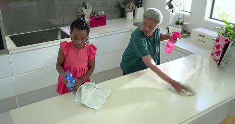 Grandmother and granddaughter cleaning bright modern kitchen countertop, family teamwork